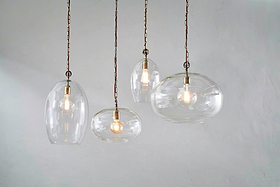Four nkuku Otoro Recycled Glass Pendants in Clear Small Oval hang at varying heights with exposed bulbs and brass fittings against a light gray wall, each elegant glass shade showcasing a glowing bulb inside.