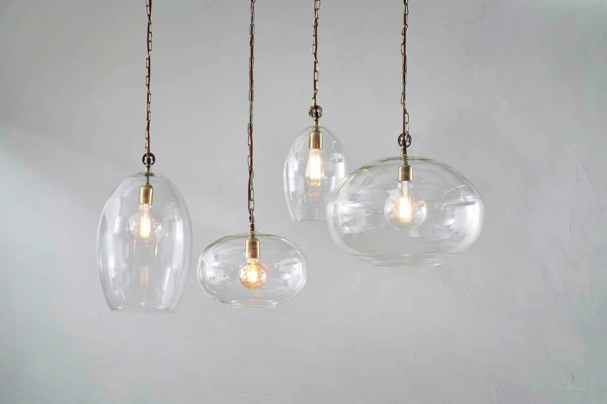 Four nkuku Otoro Recycled Glass Pendants in Clear Small Oval hang at varying heights with exposed bulbs and brass fittings against a light gray wall, each elegant glass shade showcasing a glowing bulb inside.