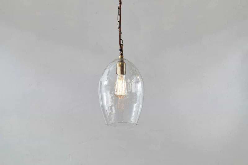 The Otoro Recycled Glass Pendant - Clear - Small Oval by nkuku features a dome-shaped glass shade and brass fittings with a vintage-style exposed bulb, making it a stylish, minimalist lighting choice.