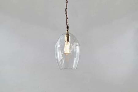 The Otoro Recycled Glass Pendant - Clear - Small Oval by nkuku features a dome-shaped glass shade and brass fittings with a vintage-style exposed bulb, making it a stylish, minimalist lighting choice.