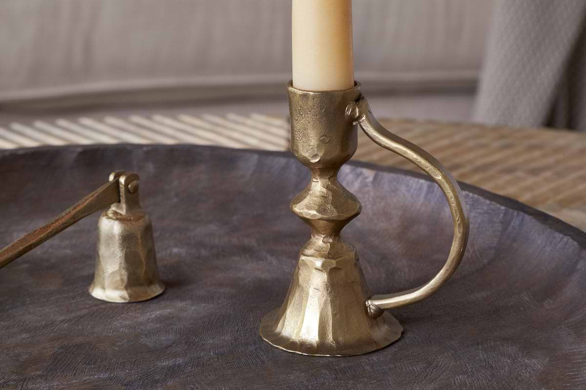 The nkuku Prema Candle Holder in antique brass, paired with a cream candle and matching snuffer, is displayed on a round metal tray against a softly blurred backdrop.
