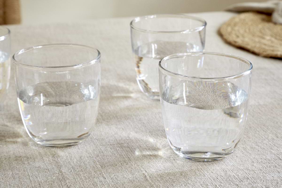 The nkuku Pani Tumbler - Clear (Set of 4) is shown filled with water on a light fabric tablecloth, with a woven placemat in the background, highlighting their lightweight and durable recycled glass design.