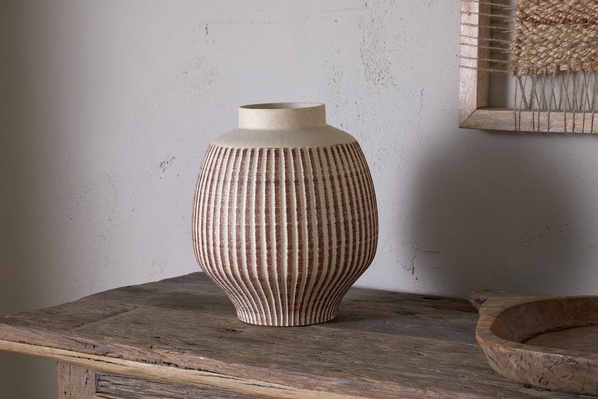 The nkuku Perola Reactive Glaze Decorative Vase in Natural & Rust sits on a rustic wooden table. Its round body tapers to a wide opening, set against a textured light gray wall with part of a framed woven artwork visible behind.