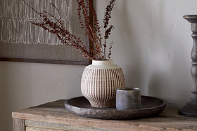 The nkuku Perola Reactive Glaze Decorative Vase in Natural & Rust holds dried brown branches on a round rustic wooden tray beside a small gray cup, with textile art hanging on a light wall in the background.