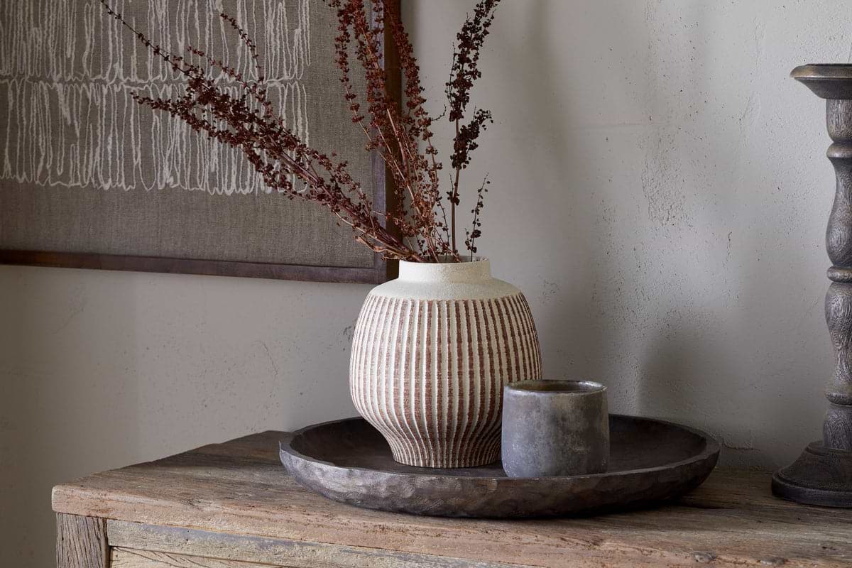 The nkuku Perola Reactive Glaze Decorative Vase in Natural & Rust holds dried brown branches on a round rustic wooden tray beside a small gray cup, with textile art hanging on a light wall in the background.