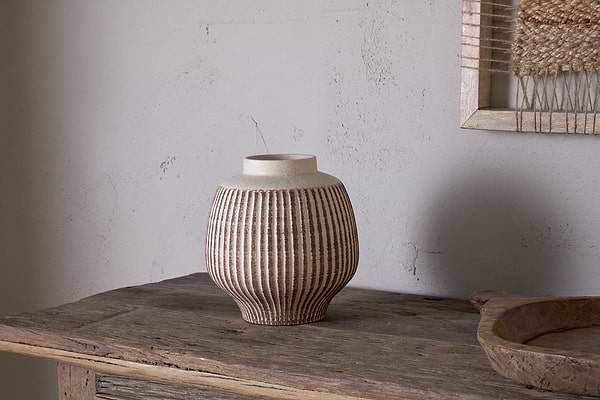 The nkuku Perola Reactive Glaze Decorative Vase in Natural & Rust, with parallel vertical brown and beige stripes, sits on a rustic wood table against a textured light wall and the edge of a framed woven artwork.