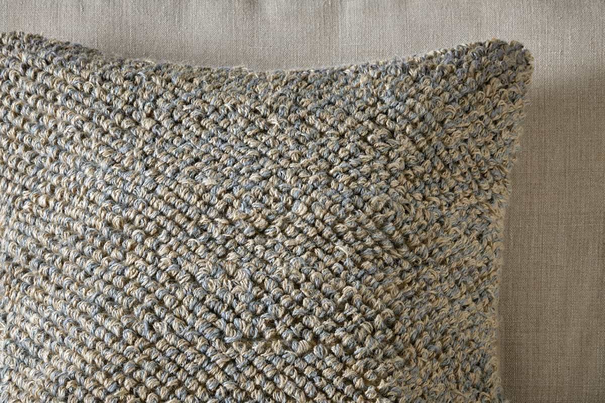 Close-up of the nkuku Paduna Jute & Cotton Cushion Cover in soft blue and brown, featuring a chunky woven texture. It rests on a taupe fabric background, its tactile look complemented by plush feather inners for added comfort.