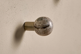 The Paladi Marble & Iron Round Hook - Grey by nkuku features a hand-carved, stone-textured marble knob in gray and beige tones, set on a short brushed brass iron base against a light beige textured wall.