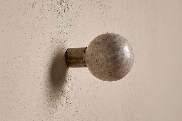The nkuku Paladi Marble & Iron Round Hook - Grey features a polished marble knob and metallic base, mounted on a textured off-white wall—its elegant, hand-carved look makes it perfect as stylish wall art hooks.