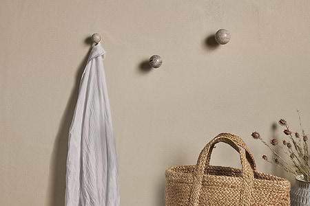 A beige wall displays three nkuku Paladi Marble & Iron Round Hooks in grey; one holds a draped white cloth. Below, a woven straw bag and vase with dried flowers rest on a surface, adding to the minimal, natural look.