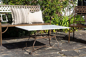 A stone patio features a wrought iron bench with two textured pillows, the nkuku Parwaha Marble Coffee Table with metal legs, and a matching chair. Green leafy plants and sunlight complete this inviting outdoor setting.