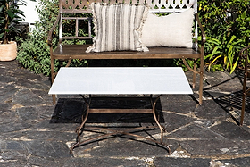 A brown metal garden bench with two striped beige pillows sits on a stone patio behind the nkuku Parwaha Marble Coffee Table with curved metal legs. Lush green potted and ground plants surround the sunlit scene.