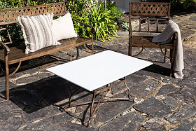 A metal bench with striped beige cushions and a matching chair draped with a gray blanket rest on a stone patio. Between them is the nkuku Parwaha Marble Coffee Table, while green plants border the patio area.