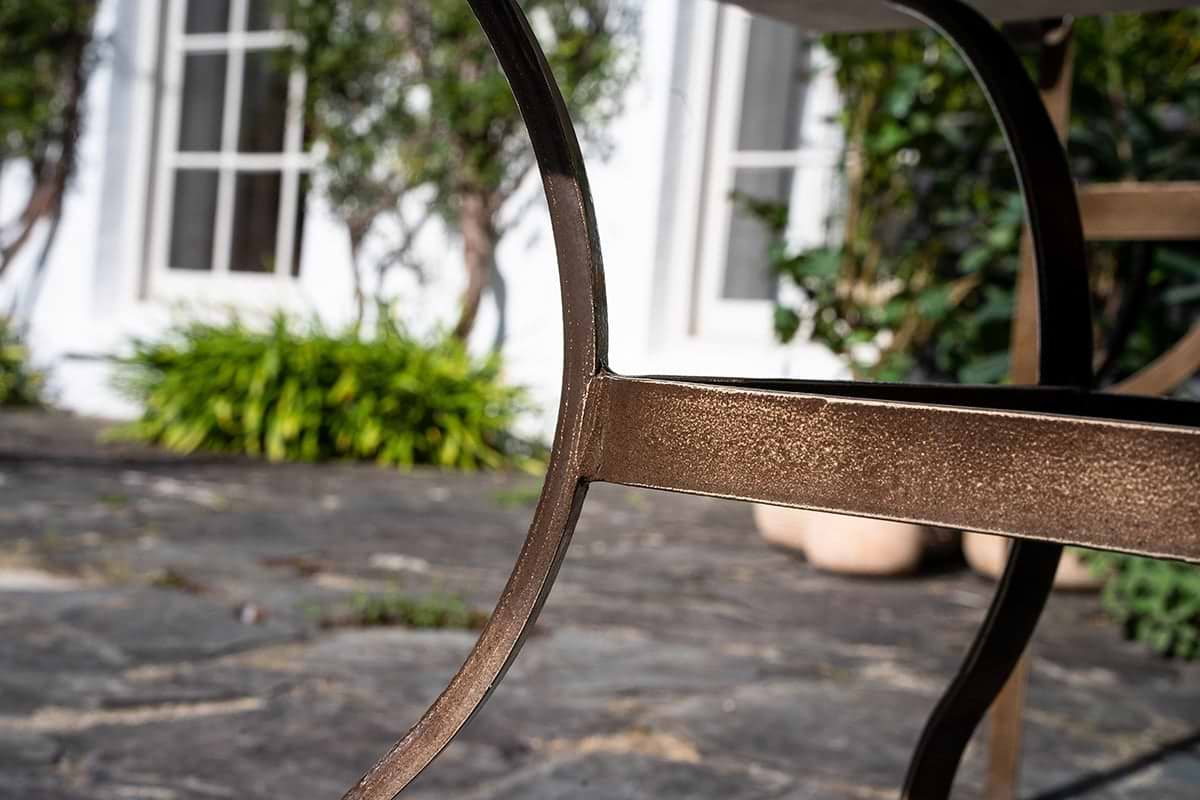Close-up of a bronze leg from the nkuku Parwaha Marble Coffee Table on a stone patio, with green plants, potted shrubs, and a white building with large windows softly blurred in the background.