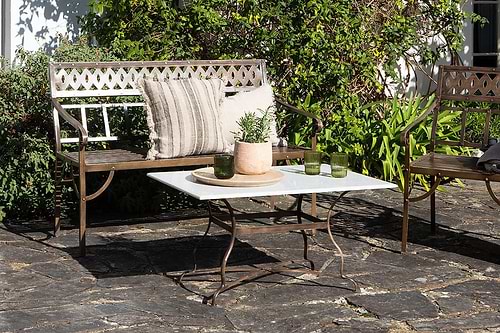A metal bench with a striped cushion and pillow sits beside the Parwaha Marble Coffee Table by nkuku, which is topped with potted plants and green glasses on a stone patio, surrounded by lush greenery in bright sunlight.