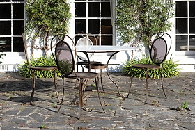 Three metal mesh-back chairs and the nkuku Parwaha Marble Dining Table are arranged on a stone patio outside a white building with large windows, bordered by green shrubs and greenery along the wall.