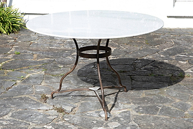The Parwaha Marble Dining Table by nkuku, featuring a round white marble top and thin curved metal legs with a circular support ring, stands on a gray stone patio. Its shadow falls nearby, with green plants visible in the upper left background.