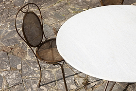 The nkuku Parwaha Marble Dining Table, with its round white marble top, sits on a stone-tiled patio beside a brown metal chair featuring a mesh seat and decorative loop backrest. Gentle sunlight completes the tranquil outdoor setting.