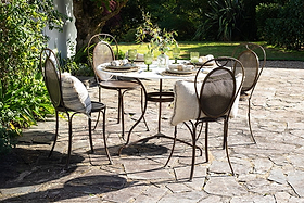 The nkuku Parwaha Marble Dining Table with four matching chairs is arranged for a meal on a stone patio, complete with plates, glasses, napkins, and chair cushions. Sunlight highlights the lush garden surrounding this inviting setup.