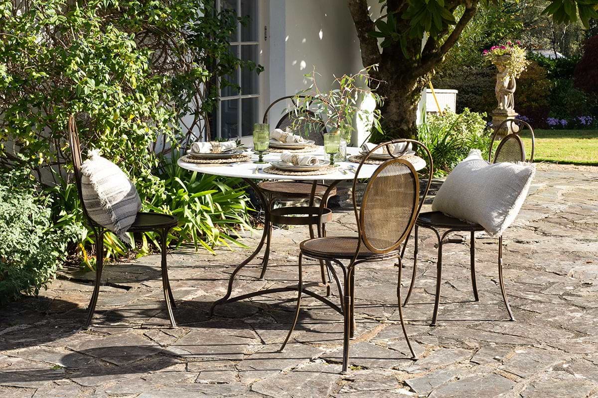 The nkuku Parwaha Marble Dining Table, set for four with cushioned metal chairs, sits on a stone patio surrounded by lush greenery and flowers, with glassware and plates atop the table and a white building in the background.
