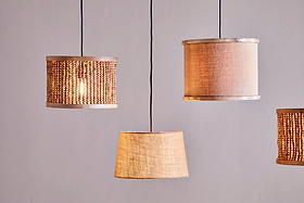 Four pendant lights with nkuku's Dia Jute Lampshade in natural hang from black cords against a light grey wall. Each shade features woven jute and wooden beads, adding textured, cozy ambient lighting.