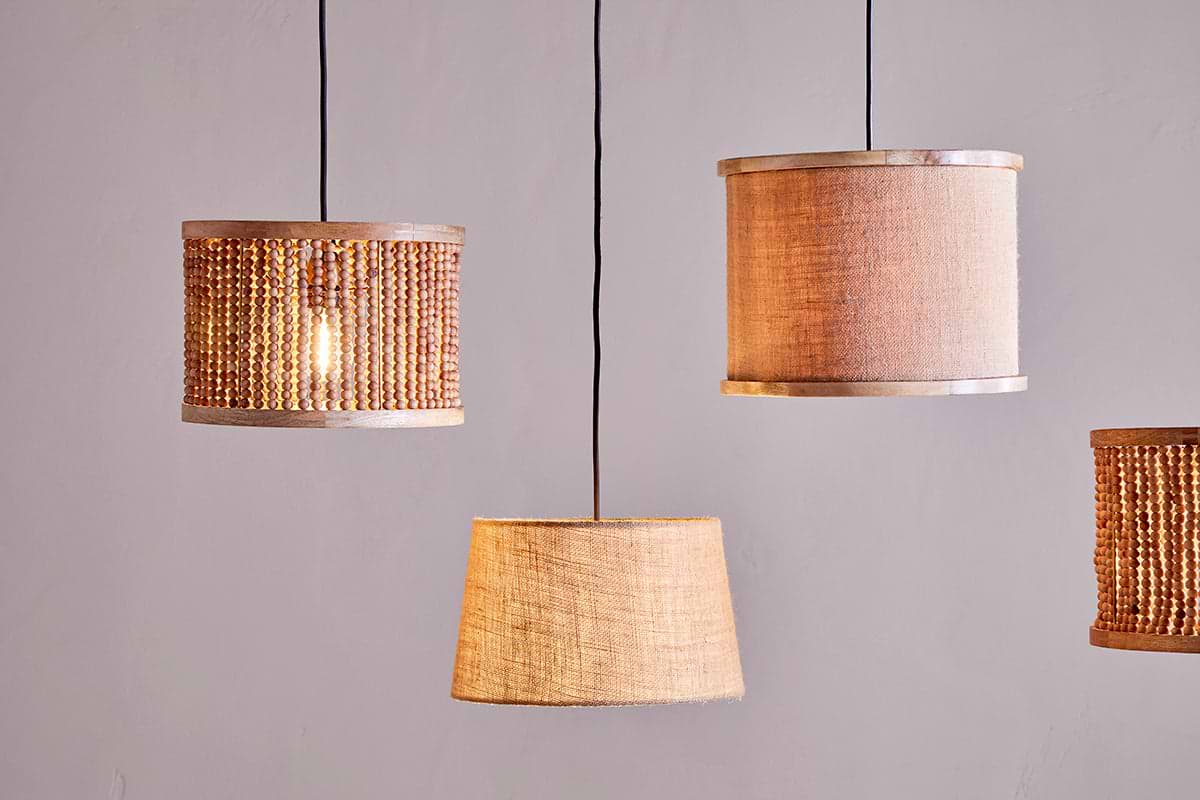 Four pendant lights with nkuku's Dia Jute Lampshade in natural hang from black cords against a light grey wall. Each shade features woven jute and wooden beads, adding textured, cozy ambient lighting.