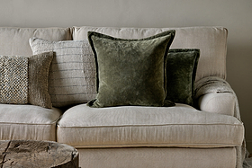 A beige fabric sofa with feather cushions features three decorative pillows: one textured beige, one light gray with thin stripes, and two nkuku Pilani Velvet & Linen Cushion Covers in Moss. A rustic wooden table sits in the front left corner against a light gray wall.