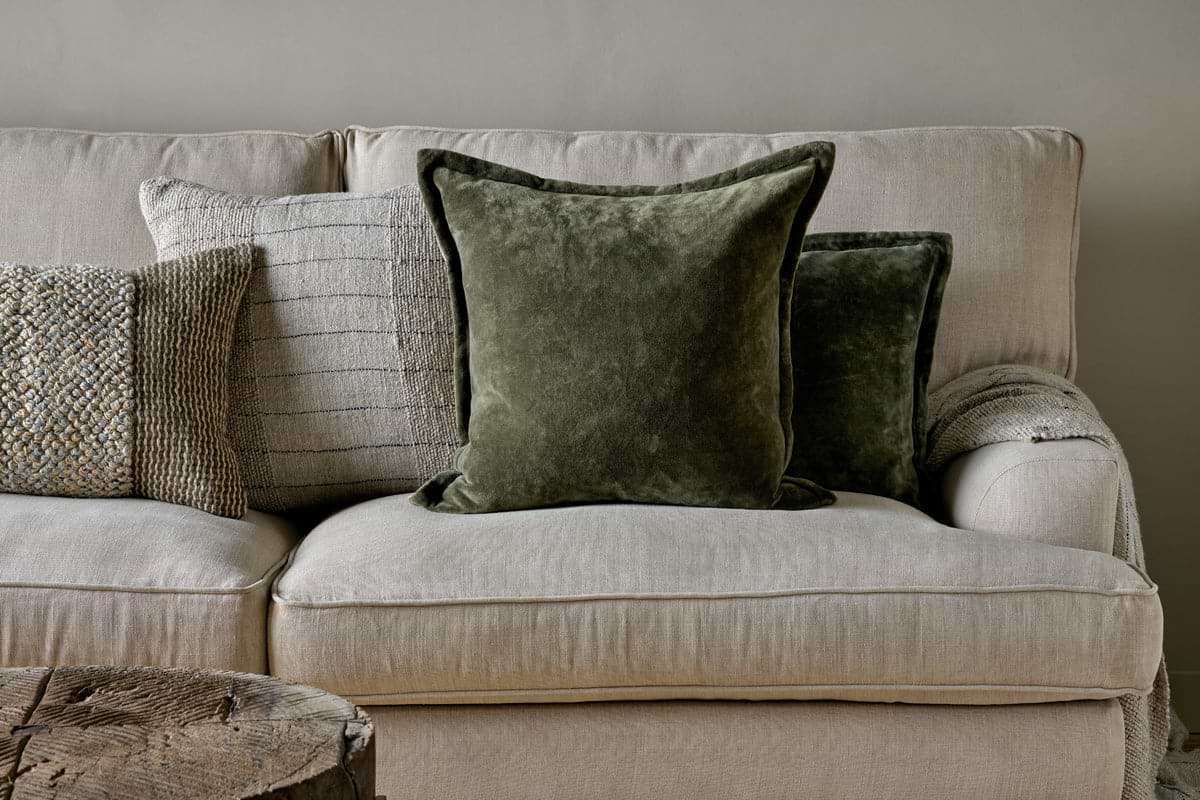 A beige fabric sofa with feather cushions features three decorative pillows: one textured beige, one light gray with thin stripes, and two nkuku Pilani Velvet & Linen Cushion Covers in Moss. A rustic wooden table sits in the front left corner against a light gray wall.
