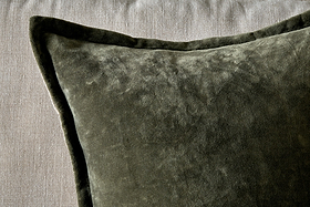 A close-up of the nkuku Pilani Velvet & Linen Cushion Cover in Moss, showcasing its soft, plush velvet texture and piped edges as it rests on a light gray fabric surface with lighting highlighting its subtle sheen.