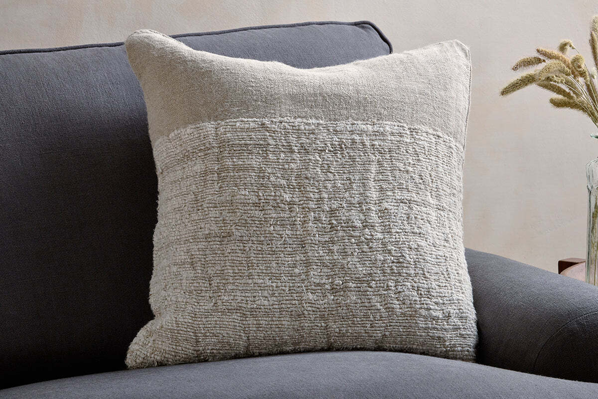 The Puli Recycled Linen Cushion Cover - Natural by nkuku, featuring a textured, natural finish, rests on a dark gray sofa. In the background, a glass vase with dried wheat stalks sits on a wooden surface for a decorative accent.