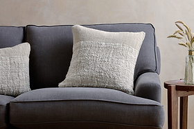A dark gray upholstered sofa features two nkuku Puli Recycled Linen Cushion Covers in Natural. Next to it stands a wooden side table with a glass vase of dried grasses, set against a subtly textured, light-colored wall.