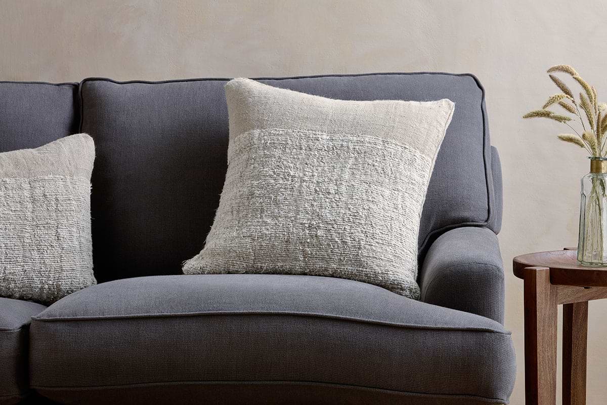 A dark gray upholstered sofa features two nkuku Puli Recycled Linen Cushion Covers in Natural. Next to it stands a wooden side table with a glass vase of dried grasses, set against a subtly textured, light-colored wall.