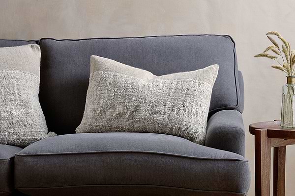 A dark gray fabric sofa with two large nkuku Puli Recycled Linen Cushion Covers in natural sits beside a small wooden side table topped with a glass vase of dried grasses, set against a light beige wall.