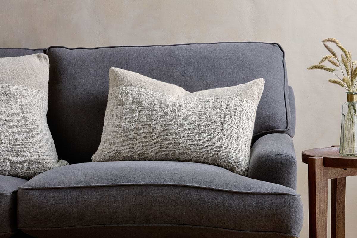 A dark gray fabric sofa with two large nkuku Puli Recycled Linen Cushion Covers in natural sits beside a small wooden side table topped with a glass vase of dried grasses, set against a light beige wall.