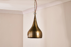The nkuku Quinn Hammered Metal Pendant – Small features a handmade, hammered teardrop shade in antique brass, suspended by a chain and leather cord, casting subtle light on beige walls and ceiling.