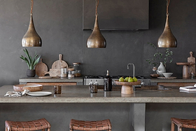 A modern kitchen features a concrete countertop, dark walls, and two woven barstools at the island. Above hang three nkuku Quinn Hammered Metal Pendants (Small, antique brass), with wood cutting boards, dishes, and pears as warm accents.