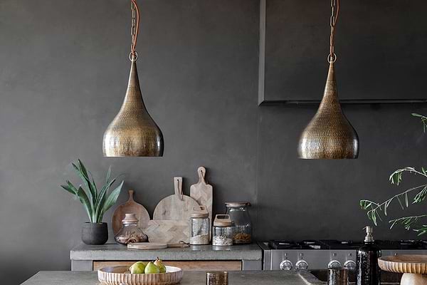 A modern kitchen features two nkuku Quinn Hammered Metal Pendant - Small lights with an antique brass finish above a gray countertop, decorated with pears on a wooden tray, a plant in a dark pot, cutting boards, jars, and utensils against a dark gray wall.