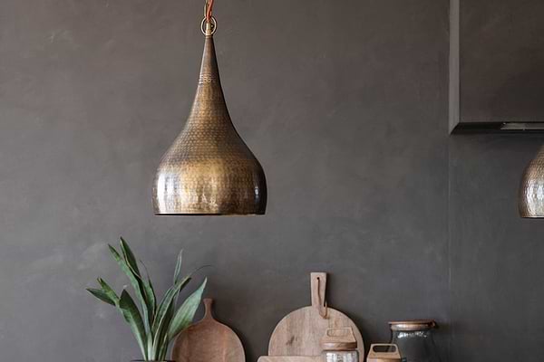 A Quinn Hammered Metal Pendant - Small by nkuku with antique brass finish and hammered texture hangs before a dark gray wall, illuminating green plant leaves, wooden cutting boards, and ceramic jars in a rustic kitchen scene.