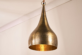 The Quinn Hammered Metal Pendant - Small by nkuku, with its teardrop shape and antique brass finish, features a handmade hammered surface that glows warmly as it hangs by a ring and wire against plain beige walls and ceiling.