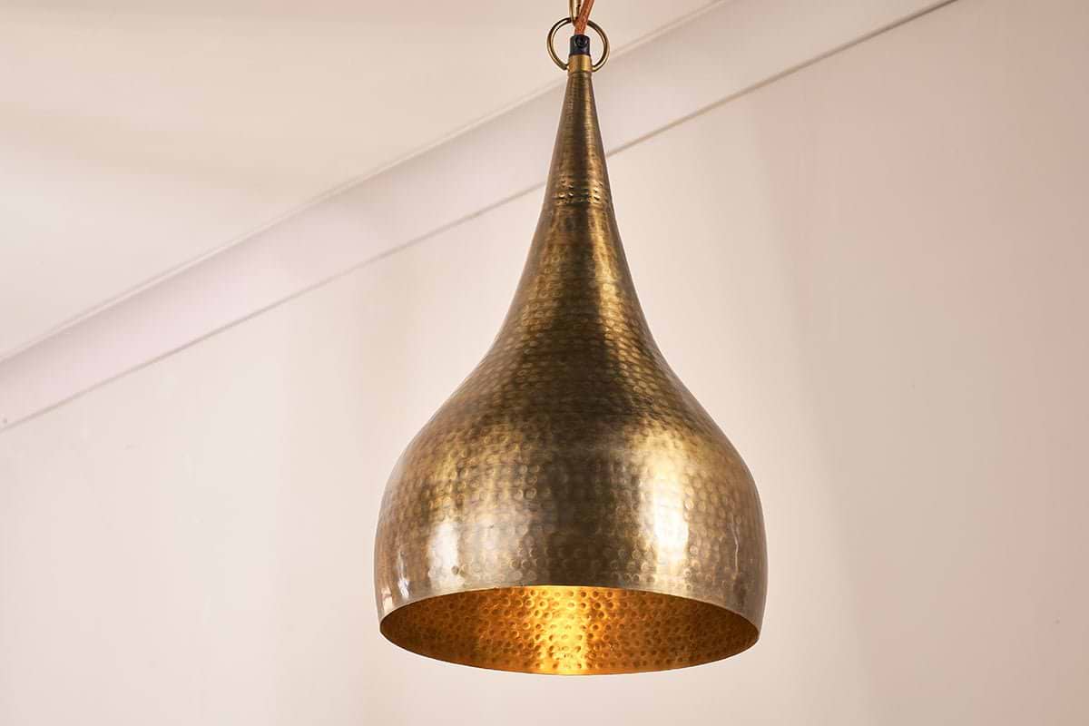 The Quinn Hammered Metal Pendant - Small by nkuku, with its teardrop shape and antique brass finish, features a handmade hammered surface that glows warmly as it hangs by a ring and wire against plain beige walls and ceiling.