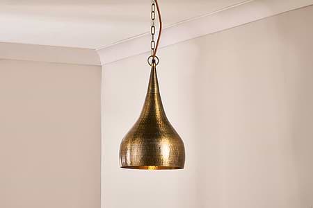 The Quinn Hammered Metal Pendant - Small by nkuku, with an antique brass hammered finish and teardrop form, hangs from a metal chain and glows warmly against the neutral beige walls and crisp white crown molding in the corner.