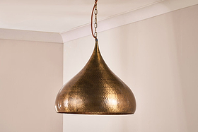 The Quinn Statement Hammered Metal Pendant - Large by nkuku, featuring a hammered metal teardrop shape, hangs from a chain in the corner of a cream-walled room with white crown molding.