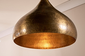 The nkuku Quinn Statement Hammered Metal Pendant - Large hangs from the ceiling, its dome-shaped, hammered surface glowing warmly and reflecting soft light against a plain off-white wall with ceiling molding.
