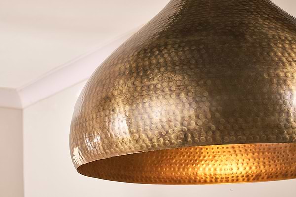 Close-up of the Quinn Statement Hammered Metal Pendant - Large by nkuku. This dome-shaped light features a hand-hammered antique brass finish, its textured surface casting a warm, golden glow throughout the room.