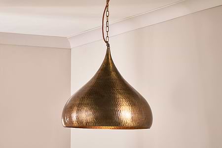 The Quinn Statement Hammered Metal Pendant - Large by nkuku hangs from a chain in the corner of a light beige room. Finished in antique brass, its teardrop hammered design casts an inviting, soft glow with textured elegance.