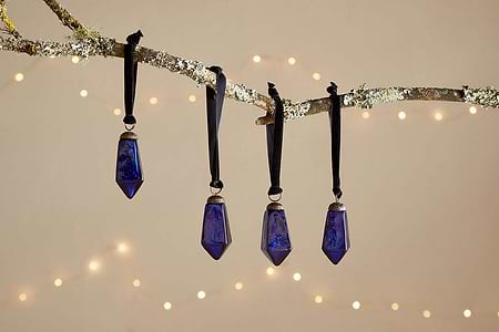 Four small, faceted indigo Ratnam Baubles by nkuku hang from black ribbons on a lichen-covered branch. Warm white fairy lights glow against a soft beige background, creating a festive, cozy scene. Set of 4 included.