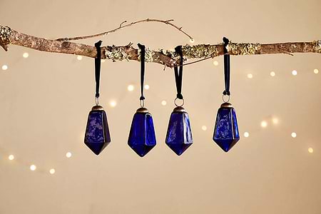 Four Ratnam Baubles - Indigo - Large (Set of 4) by nkuku, featuring geometric shapes and an antique glass finish, hang from black ribbons on a lichen branch. Soft fairy lights glow behind, evoking classic Christmas charm.