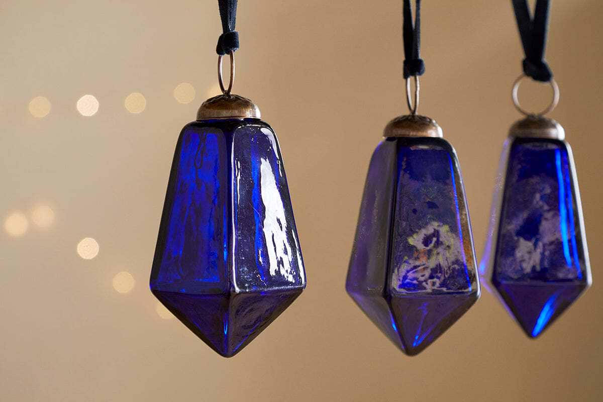 The Ratnam Baubles - Indigo - Large (Set of 4) by nkuku feature deep indigo glass with varied geometric patterns and metallic tops, hanging from black cords to create a warm, elegant look against a soft beige background with blurred lights.
