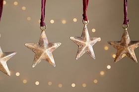 The nkuku Roshani Star Baubles - Antique Brass (Set of 4) hang from red strings against a beige backdrop with soft string lights. Only parts of the two outer star silhouettes are visible at the edges, creating a warm, festive feel.