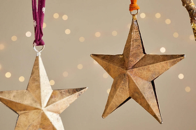 The nkuku Roshani Star Baubles - Antique Brass (Set of 2) feature metallic gold stars hanging from purple and orange ribbons, set against a soft beige backdrop with blurred lights for a festive, warm holiday ambiance.